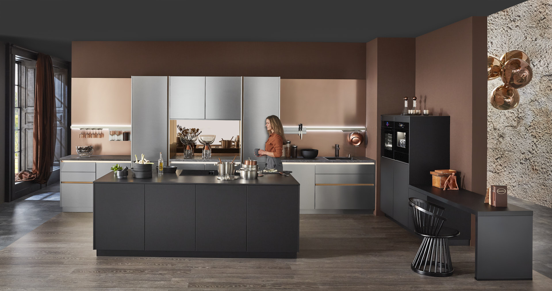 Hauptschuss, Mix aus Keramik und Bronzenachbildung, Kücheninsel mit Bosch Induktionskochfeld und Muldenlüfter, schwarzer Schrank, Edelstahlfronten, Bronze Nischenrückwand, 2 Bosch Einbaubacköfen, Messing Spüle, Model ist hinter Kücheninsel